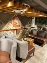 The image features a cozy indoor seating area with modern gray and brown chairs arranged around a small table. A part of the wall is adorned with a large, colorful mural depicting people in a playful pose. The ceiling is detailed with wooden paneling and modern lighting. A cardboard box is placed on the floor next to the seating area, and there is some greenery visible on the wall to the right.