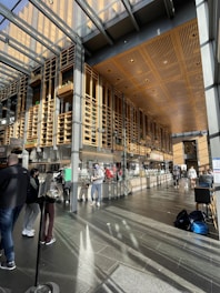 A modern food court area with wooden and metal architectural elements. There are people casually interacting and purchasing food from the vendors lined up along the wall. Sunlight streams through large glass panels in the ceiling, casting patterns on the floor. Backpacks and seating arrangements are visible, contributing to the busy yet organized atmosphere.