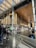 A modern food court area with wooden and metal architectural elements. There are people casually interacting and purchasing food from the vendors lined up along the wall. Sunlight streams through large glass panels in the ceiling, casting patterns on the floor. Backpacks and seating arrangements are visible, contributing to the busy yet organized atmosphere.