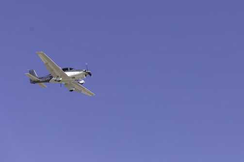 A light aircraft with a propeller is flying at a significant altitude against a clear blue sky. The plane has a sleek design and the registration number 'N323KG' is visible on its body.