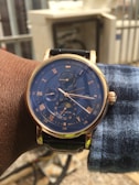 A wristwatch with a blue face and gold-tone casing is shown. The watch features multiple subdials, labeled with Roman numerals and various indicators such as a tachymeter and moon phase. The dial is detailed with minute and hour markers, and the band appears to be black leather. The watch is worn on the left wrist of a person with dark skin, and they are wearing a blue and white checkered shirt. The background is blurred but shows some outdoor elements, including a fence and some building structures.