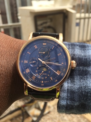 A wristwatch with a blue face and gold-tone casing is shown. The watch features multiple subdials, labeled with Roman numerals and various indicators such as a tachymeter and moon phase. The dial is detailed with minute and hour markers, and the band appears to be black leather. The watch is worn on the left wrist of a person with dark skin, and they are wearing a blue and white checkered shirt. The background is blurred but shows some outdoor elements, including a fence and some building structures.