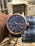 A wristwatch with a blue face and gold-tone casing is shown. The watch features multiple subdials, labeled with Roman numerals and various indicators such as a tachymeter and moon phase. The dial is detailed with minute and hour markers, and the band appears to be black leather. The watch is worn on the left wrist of a person with dark skin, and they are wearing a blue and white checkered shirt. The background is blurred but shows some outdoor elements, including a fence and some building structures.