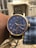 A wristwatch with a blue face and gold-tone casing is shown. The watch features multiple subdials, labeled with Roman numerals and various indicators such as a tachymeter and moon phase. The dial is detailed with minute and hour markers, and the band appears to be black leather. The watch is worn on the left wrist of a person with dark skin, and they are wearing a blue and white checkered shirt. The background is blurred but shows some outdoor elements, including a fence and some building structures.