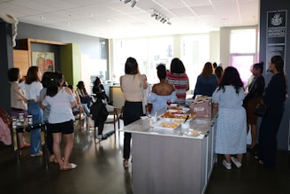 A group of people is gathered in a modern interior space, possibly a cafe or a meeting area. They are casually dressed and standing around a table with food and beverages, suggesting a social or networking event. The room is well-lit with large windows in the background and features contemporary decor.