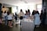 A group of people is gathered in a modern interior space, possibly a cafe or a meeting area. They are casually dressed and standing around a table with food and beverages, suggesting a social or networking event. The room is well-lit with large windows in the background and features contemporary decor.