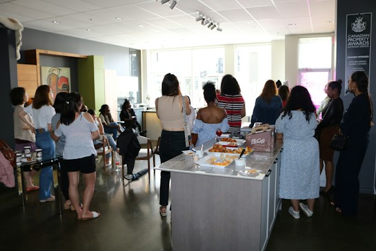 A group of people is gathered in a modern interior space, possibly a cafe or a meeting area. They are casually dressed and standing around a table with food and beverages, suggesting a social or networking event. The room is well-lit with large windows in the background and features contemporary decor.