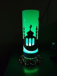 A cylindrical lamp emits a soft green glow, featuring intricate cut-out designs of a mosque with minarets and a crescent moon. The base of the lamp is ornately decorated with golden details, enhancing its elegant appearance.