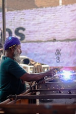 An older man with a beard and a purple headscarf is engaged in welding, with bright sparks emanating from the welding point. He is focused on his work, holding a welding tool in his right hand, with protective gear visible on his ear. The background is an outdoor setting with a wall that has faded writing on it.