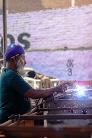 An older man with a beard and a purple headscarf is engaged in welding, with bright sparks emanating from the welding point. He is focused on his work, holding a welding tool in his right hand, with protective gear visible on his ear. The background is an outdoor setting with a wall that has faded writing on it.