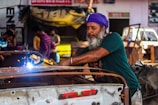A man wearing a purple headscarf and green shirt is welding metal in a garage. Sparks fly from the welding tool he holds. Other people and various car parts are visible in the background, creating a bustling workshop atmosphere.