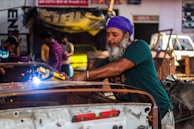 A man wearing a purple headscarf and green shirt is welding metal in a garage. Sparks fly from the welding tool he holds. Other people and various car parts are visible in the background, creating a bustling workshop atmosphere.
