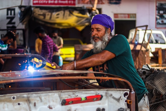 A man wearing a purple headscarf and green shirt is welding metal in a garage. Sparks fly from the welding tool he holds. Other people and various car parts are visible in the background, creating a bustling workshop atmosphere.