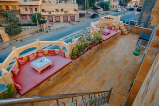 A rooftop terrace features a cozy seating area with red cushions surrounding a small table with a decorative cloth. Potted plants line the edge of the terrace, adding greenery and contrast to the stone-colored flooring. The space overlooks a quiet street lined with buildings, some of which have greenery on their walls.