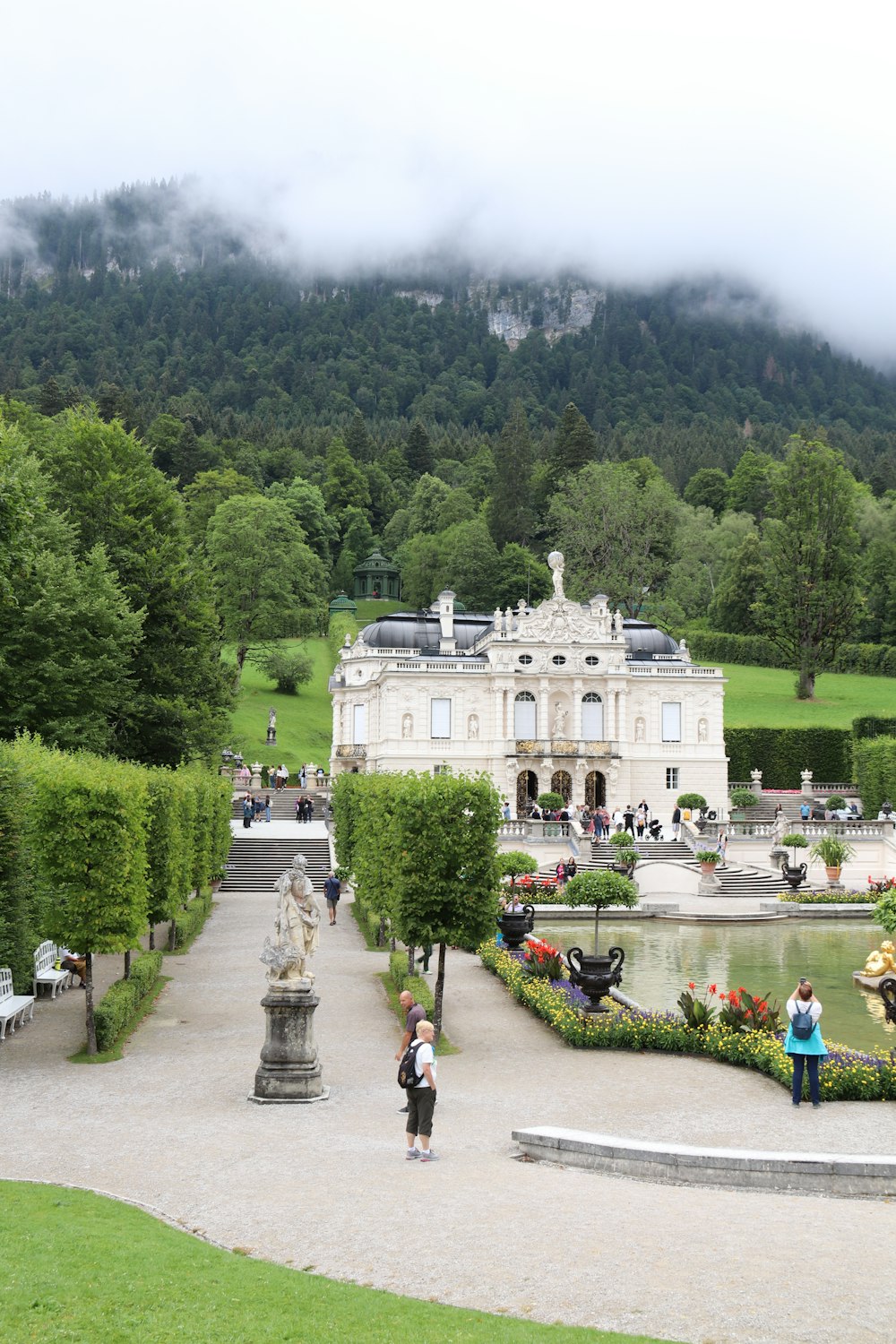 a large white building with a fountain in front of it and people walking around with Linderhof Palace in the background