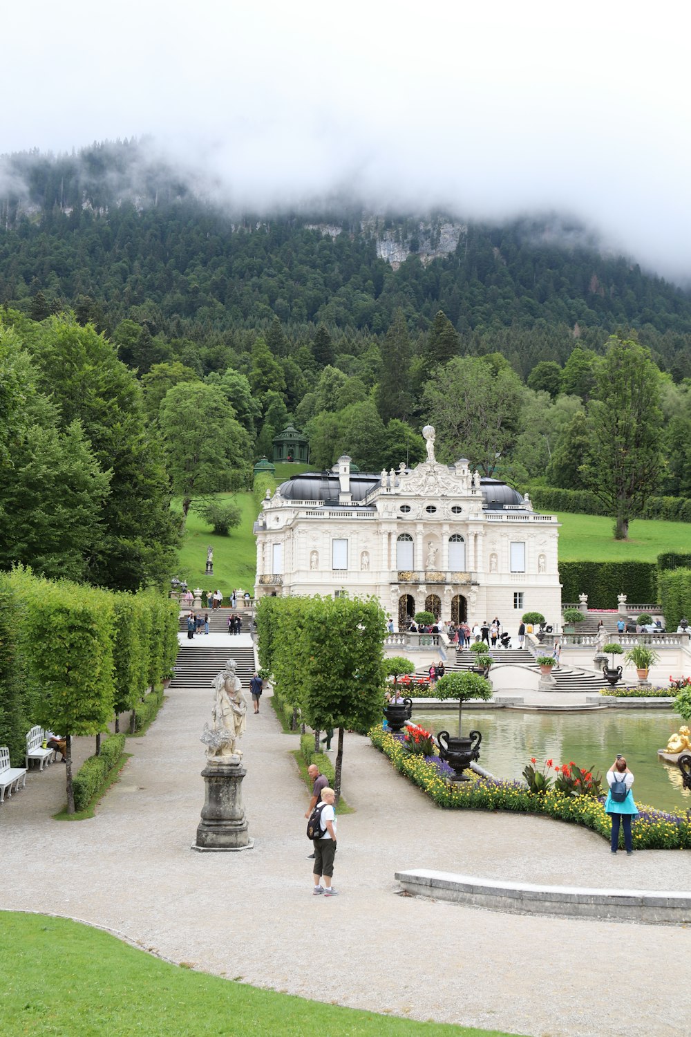 ein großes weißes Gebäude mit einem Brunnen davor und Menschen, die mit Schloss Linderhof im Hintergrund herumlaufen