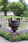 A meticulously landscaped garden featuring a large decorative urn with intricate designs, holding a neatly trimmed tree. It is set against a backdrop of manicured lawns, vibrant flowerbeds, and a tranquil pond. Various flowering plants, including purple and yellow blooms, accentuate the scene.