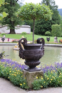 A meticulously landscaped garden featuring a large decorative urn with intricate designs, holding a neatly trimmed tree. It is set against a backdrop of manicured lawns, vibrant flowerbeds, and a tranquil pond. Various flowering plants, including purple and yellow blooms, accentuate the scene.