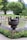 A meticulously landscaped garden featuring a large decorative urn with intricate designs, holding a neatly trimmed tree. It is set against a backdrop of manicured lawns, vibrant flowerbeds, and a tranquil pond. Various flowering plants, including purple and yellow blooms, accentuate the scene.