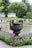 A meticulously landscaped garden featuring a large decorative urn with intricate designs, holding a neatly trimmed tree. It is set against a backdrop of manicured lawns, vibrant flowerbeds, and a tranquil pond. Various flowering plants, including purple and yellow blooms, accentuate the scene.
