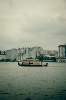 A traditional wooden houseboat is floating on a large body of water, with a backdrop of modern high-rise residential buildings under an overcast sky.