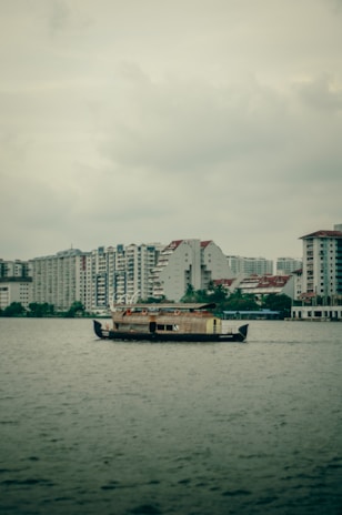 A traditional wooden houseboat is floating on a large body of water, with a backdrop of modern high-rise residential buildings under an overcast sky.