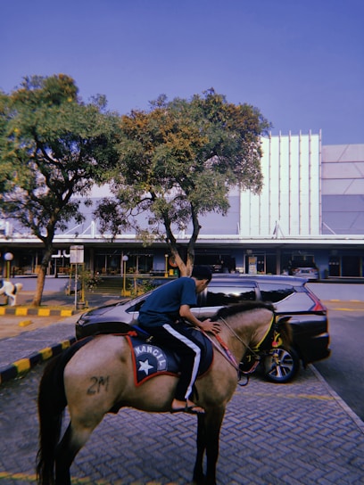 A person wearing casual clothes is riding a horse with a saddle marked with a star and the text 'KANCIL'. The scene takes place in an urban setting with a parked car nearby and buildings in the background. Trees with green foliage add a touch of nature to the environment.