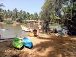 A serene riverside scene with several people engaged in boating activities. A houseboat is docked near the shore, and two empty kayaks are visible on the sandy bank. The area is lush with greenery, including overhanging trees and a backdrop of palm trees across the river. The setting conveys a peaceful, outdoor recreational atmosphere.
