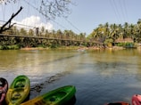 A serene river with a suspension bridge spans across the water. Colorful kayaks are docked along the riverbank in the foreground, while palm trees and a small hut line the opposite shore. Several people are seen enjoying activities on the river, creating an atmosphere of leisure and tranquility.