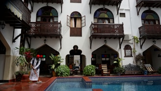 A courtyard with a pool at the center is surrounded by a traditional building with white walls and dark wooden balconies. A person wearing a traditional outfit is mopping the floor near the pool. There are lush green plants and colorful stained glass windows that add vibrancy to the scene.