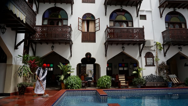 A courtyard with a pool at the center is surrounded by a traditional building with white walls and dark wooden balconies. A person wearing a traditional outfit is mopping the floor near the pool. There are lush green plants and colorful stained glass windows that add vibrancy to the scene.