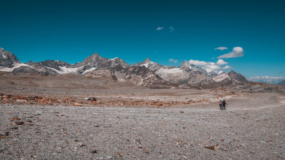 a couple people walking on a rocky terrain with mountains in the background