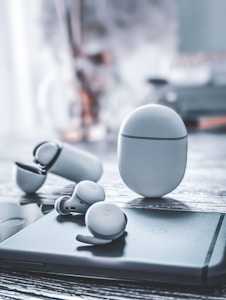 A pair of wireless earbuds and their charging case are placed on a sleek, modern surface. The design is minimalistic with a smooth, rounded shape. The backdrop is blurred, creating a focus on the earbuds.