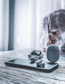 A pair of wireless earbuds and their charging case are placed on top of a smartphone, all set on a textured, gray wooden surface. In the background, a vase with some blurred flowers can be seen.