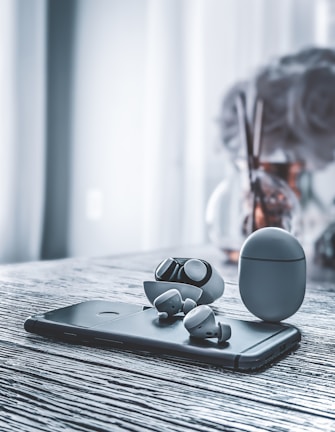 A pair of wireless earbuds and their charging case are placed on top of a smartphone, all set on a textured, gray wooden surface. In the background, a vase with some blurred flowers can be seen.
