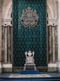 A luxurious throne room with an ornate silver throne set against an opulent green tapestry adorned with heraldic symbols. The room features classical architecture with marble columns and archways, creating a regal atmosphere.