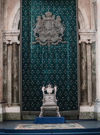 A luxurious throne room with an ornate silver throne set against an opulent green tapestry adorned with heraldic symbols. The room features classical architecture with marble columns and archways, creating a regal atmosphere.