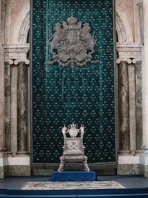 A luxurious throne room with an ornate silver throne set against an opulent green tapestry adorned with heraldic symbols. The room features classical architecture with marble columns and archways, creating a regal atmosphere.