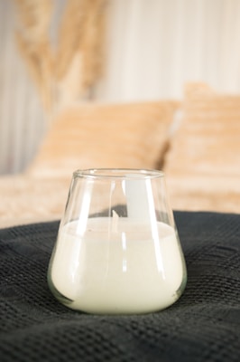 A glass candle holder containing a white candle is placed on a textured dark fabric surface. In the blurred background, there are soft-focus beige pillows and light-colored drapes or curtains, creating a cozy and elegant atmosphere.