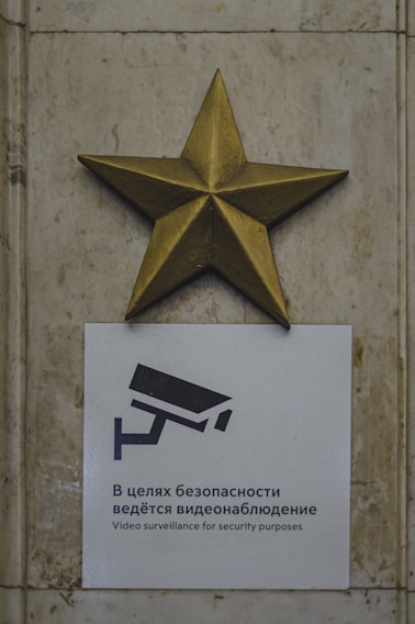 A metallic golden star is mounted above a sign featuring a camera icon and text in both Russian and English about video surveillance for security purposes. The background consists of a stone or marble wall with a textured appearance.
