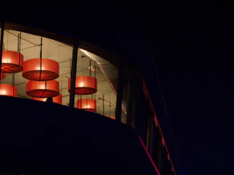 A corner of a building features a large window through which several large, circular red light fixtures are visible. The interior appears to be warmly lit, contrasting with the dark exterior surrounding the building.