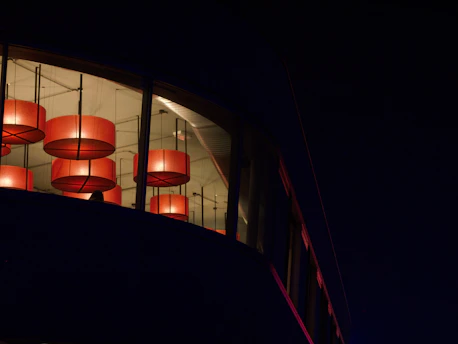 A corner of a building features a large window through which several large, circular red light fixtures are visible. The interior appears to be warmly lit, contrasting with the dark exterior surrounding the building.