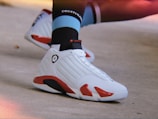 A close-up of a foot wearing a white and red athletic shoe with a jumpman logo. The shoe is paired with a black, light blue, and white sock that has text on it. The background is a neutral-colored floor, creating a casual and sporty vibe.