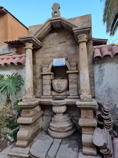 An ornate stone structure with columns and an archway, housing a central fountain or basin labeled 'Agua Bendita.' It features decorative carvings and is set against an aged stone wall with foliage and stacked roof tiles nearby.