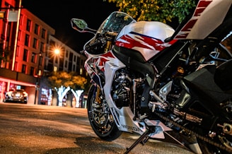 A sleek, sporty motorcycle is parked on an urban street at night. The motorcycle features a white body with bold red and black graphic designs. Nearby streetlights and buildings emit a warm glow, casting reflections and creating a vibrant nighttime scene. Trees with illuminated decorations add a festive touch to the cityscape.