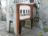A piece of urban street art depicting an old television painted on a concrete block against a textured wall. The TV screen is white with the words 'FREE BRAINWASHING' in bold black letters. Next to it is a faded image of a person's face. The art combines vibrant colors and rough textures, creating a striking visual commentary.