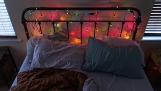 An unmade bed with a black metal headboard adorned with colorful string lights. The bed has two pillows, one light blue and one with a mix of colors, and a brown blanket. The room features two windows with blinds, creating a cozy ambience with a mix of light and shadow.
