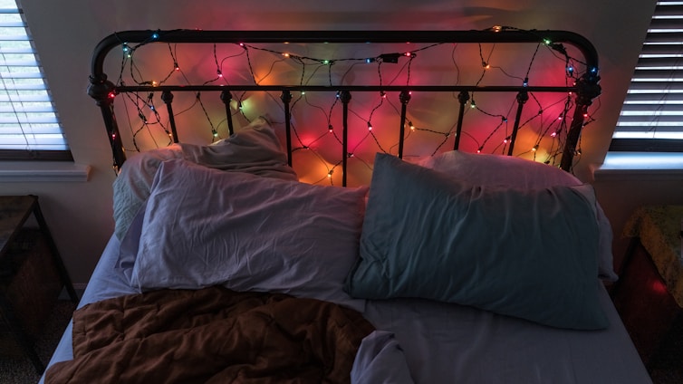 An unmade bed with a black metal headboard adorned with colorful string lights. The bed has two pillows, one light blue and one with a mix of colors, and a brown blanket. The room features two windows with blinds, creating a cozy ambience with a mix of light and shadow.
