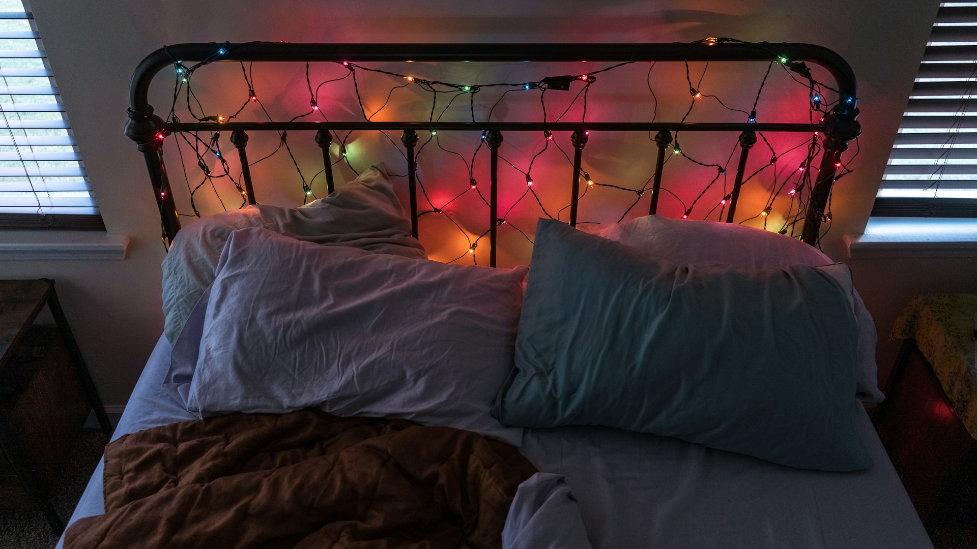 An unmade bed with a black metal headboard adorned with colorful string lights. The bed has two pillows, one light blue and one with a mix of colors, and a brown blanket. The room features two windows with blinds, creating a cozy ambience with a mix of light and shadow.