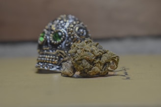 A detailed, ornamental metal skull with green gemstone eyes is positioned in the background, while a piece of dried plant material, possibly marijuana, is placed in front of it. The surface they are on appears smooth and beige, and the background is blurred.